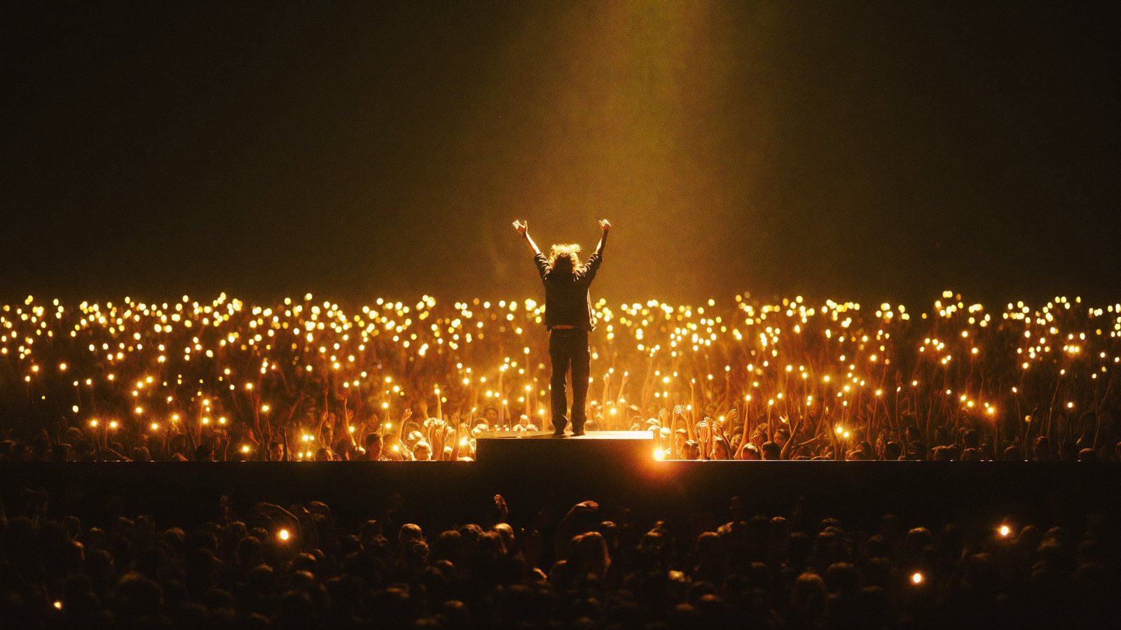 A music artist on stage, arms raised, facing a vast crowd holding up a sea of warm golden lights